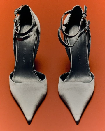 Two grey high-heeled shoes with ankle straps on a bright orange backdrop, studio shot, showcasing fashionable footwear.