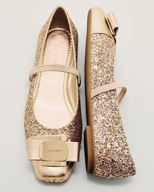 Shiny golden ballet flats with bow embellishment, one upright and one lying on its side, isolated against a light blue background.