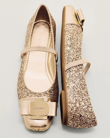 Shiny golden ballet flats with bow embellishment, one upright and one lying on its side, isolated against a light blue background.