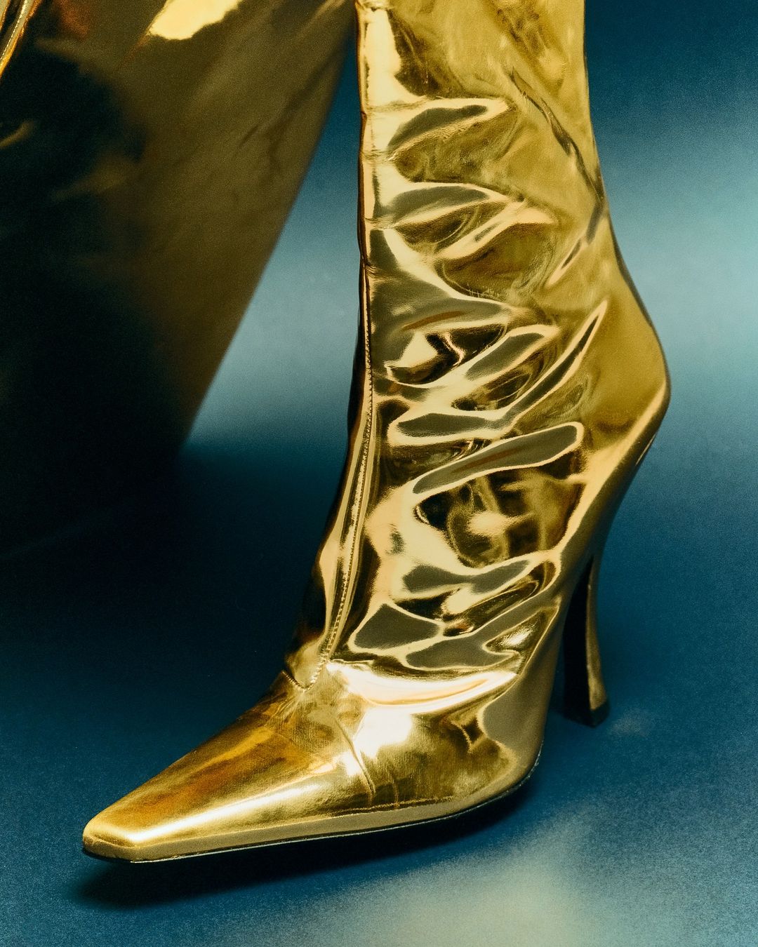 Close-up of a glossy gold high-heeled boot with a pointed toe, set against a dark teal background, showcasing fashion footwear.