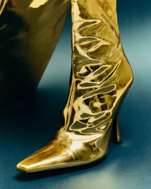 Close-up of a glossy gold high-heeled boot with a pointed toe, set against a dark teal background, showcasing fashion footwear.