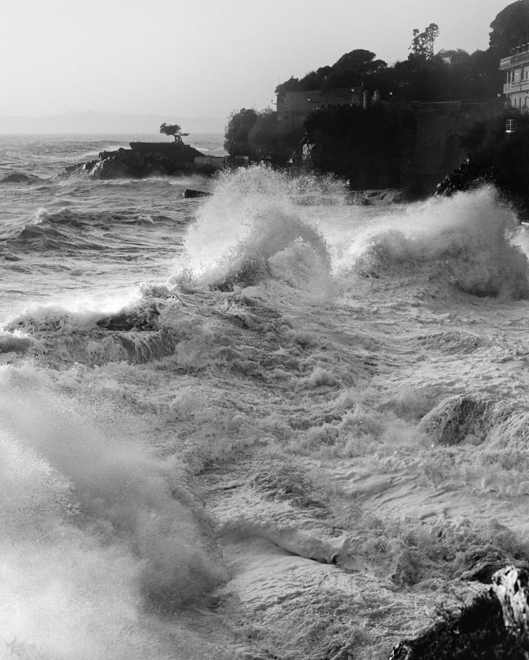 Monochrome seascape featuring large, powerful waves crashing against the shoreline, with a rocky outcrop and building visible in the background.