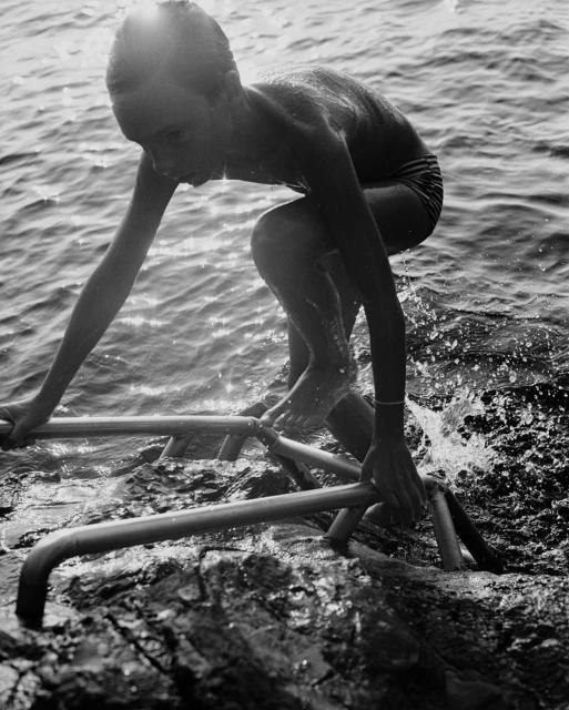 Black and white photo of a young boy in a swimsuit climbing out of the ocean onto rocks using a metal ladder.