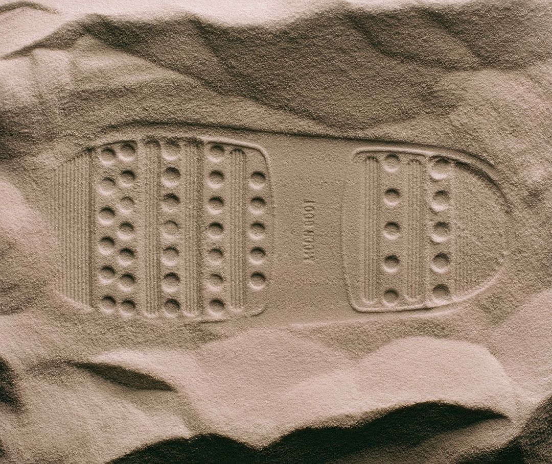 Footprint of a moon boot tread pattern in a sandy surface with a sepia tone, highlighting the textured impression in the monochromatic shot.
