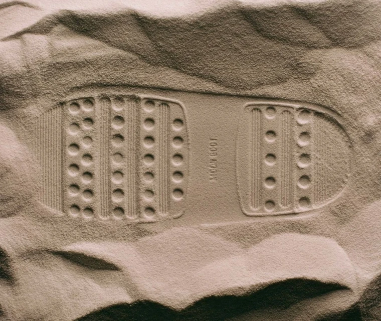 Footprint of a moon boot tread pattern in a sandy surface with a sepia tone, highlighting the textured impression in the monochromatic shot.