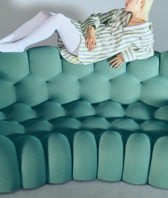 A woman relaxes on a quilted, teal-colored sofa wearing a striped pajama top and white tights, in a minimalist, indoor setting.