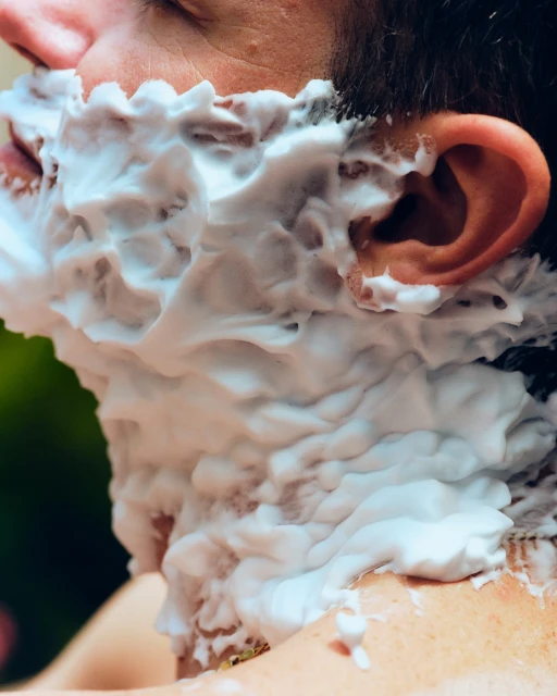 Close-up of an adult male's face covered in white shaving cream, with focus on texture and skin details, shot in bright outdoor light.
