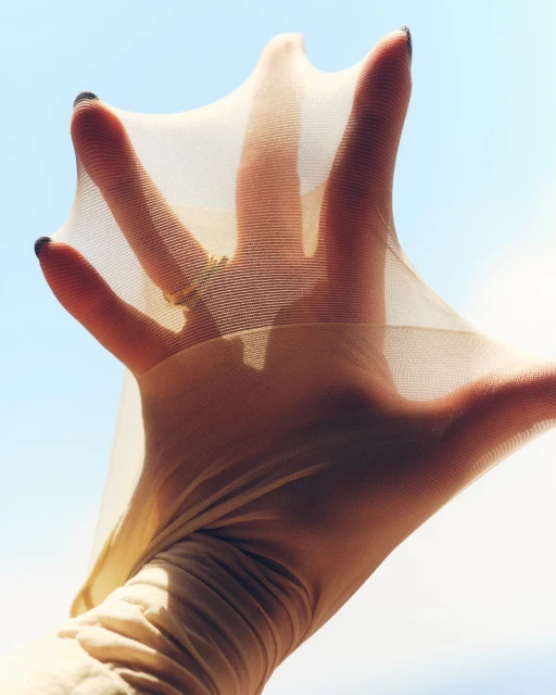 Close-up of an adult hand stretching a sheer, nude-colored stocking against a clear, bright blue sky, showing the weave of the fabric.
