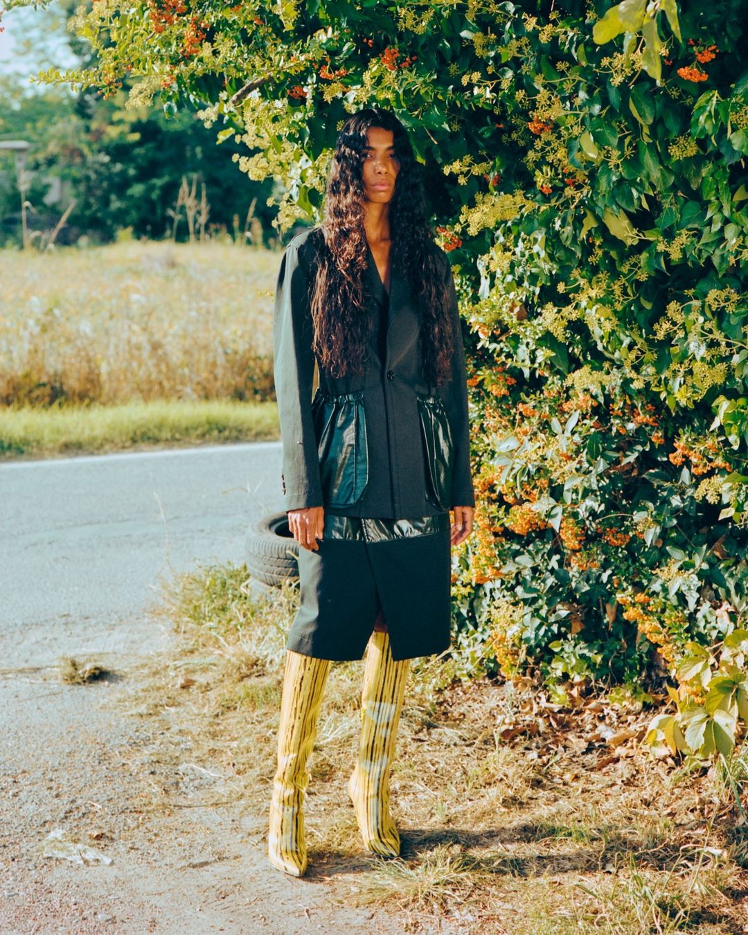 Fashionable woman poses in blazer and boots on a rural road, greenery and vegetation in background, creating a striking outdoor fashion shot.
