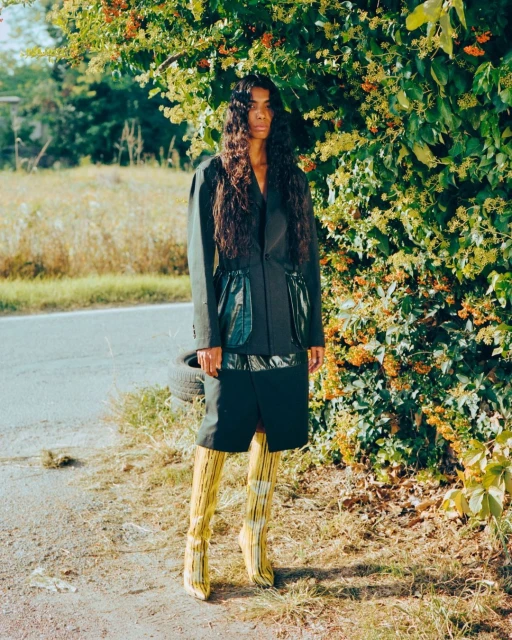 Fashionable woman poses in blazer and boots on a rural road, greenery and vegetation in background, creating a striking outdoor fashion shot.