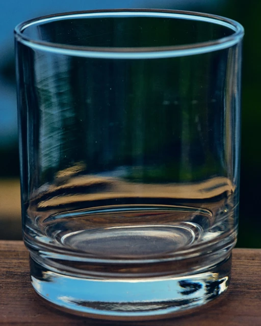 A clear, empty glass tumbler sits on a wooden surface with a blurry green and blue background, creating a simple yet elegant still life.