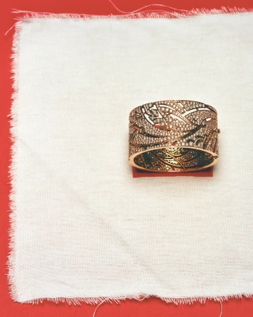 Close-up of an ornate rose gold and diamond bangle bracelet on a white linen cloth atop a red background, showcasing its intricate design.