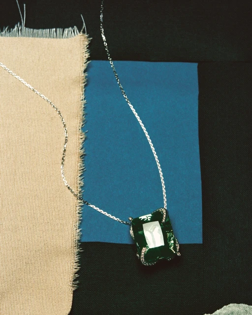 Elegant necklace with silver chain and large emerald-colored gemstone pendant on tan and blue textured fabric background, jewelry still life.