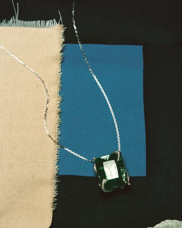 Elegant necklace with silver chain and large emerald-colored gemstone pendant on tan and blue textured fabric background, jewelry still life.