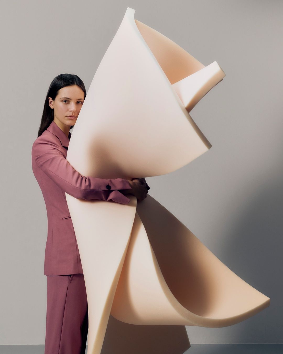 Brunette woman in suit embraces a cream-colored abstract sculptural form in a minimalist studio setting with soft, diffused lighting.