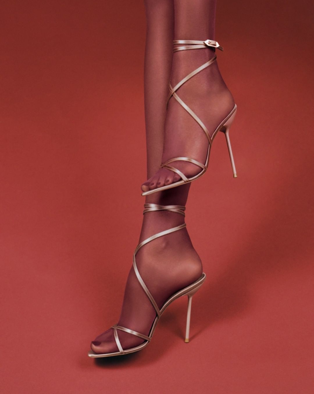 Woman's legs and feet wearing beige strappy high heels with hosiery against a red background in a studio fashion shot. 
