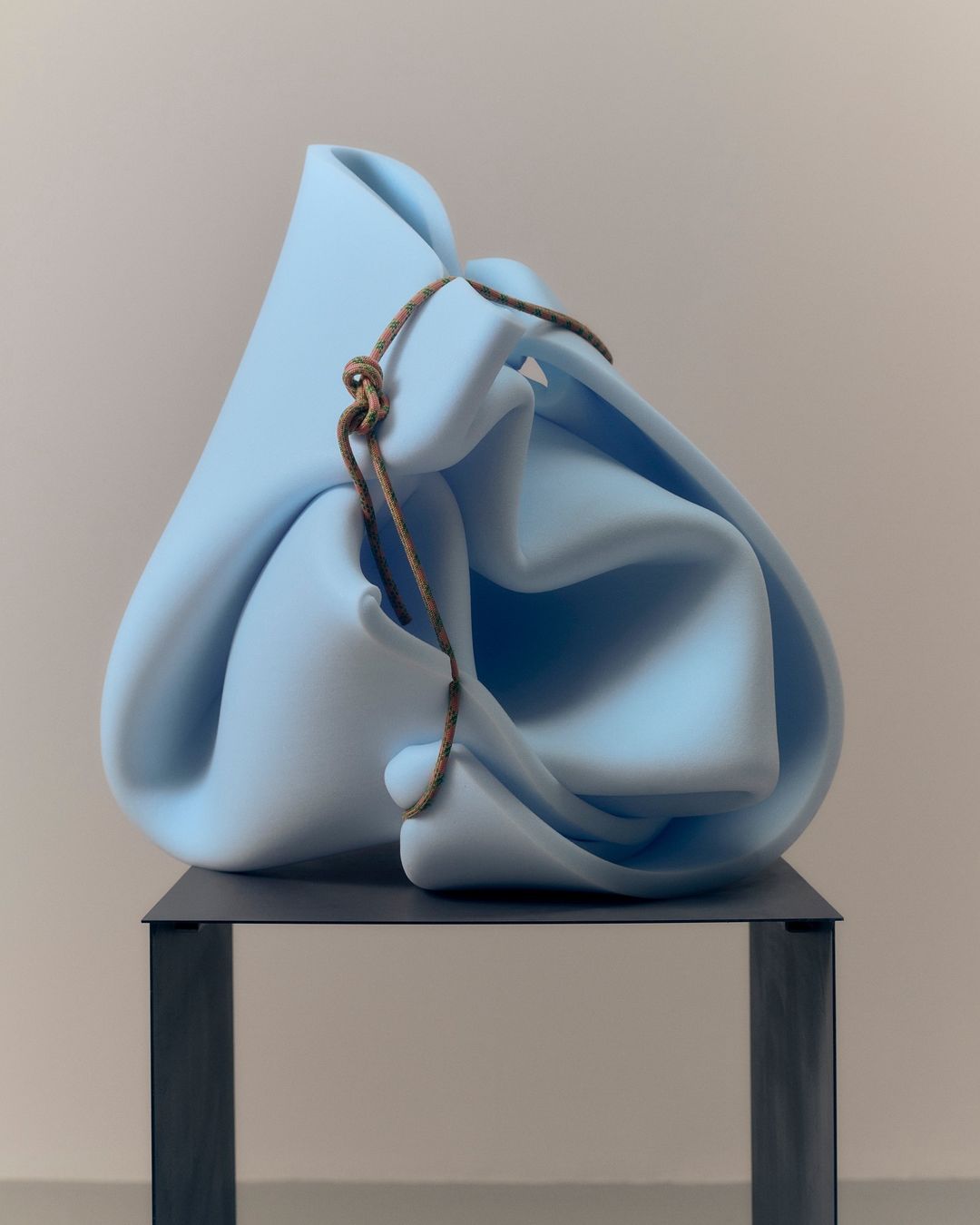 Abstract sculpture featuring a light blue, draped form on a dark gray metal stand, captured in a studio setting with soft lighting.