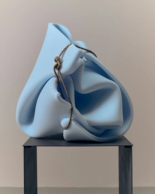 Abstract sculpture featuring a light blue, draped form on a dark gray metal stand, captured in a studio setting with soft lighting.