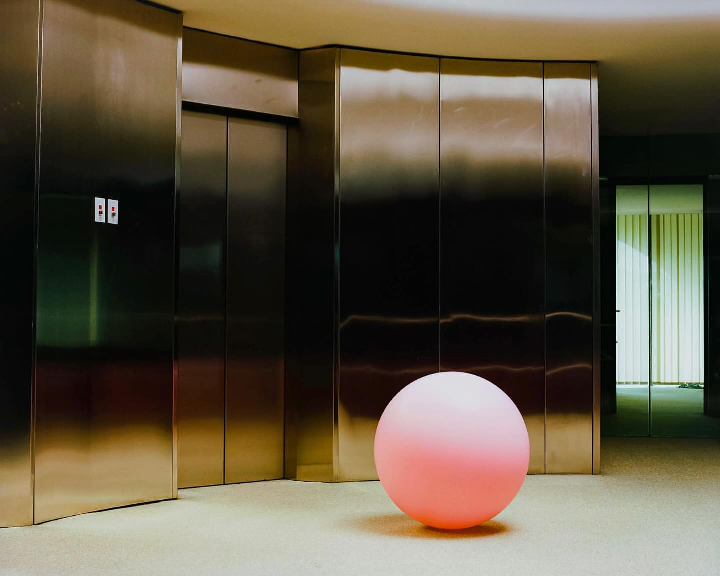 Pink sphere in a modern lobby with a carpeted floor, stainless steel elevator doors, and a glass wall with blinds.