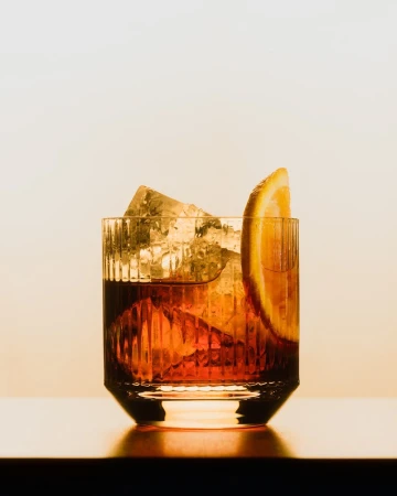 A negroni cocktail in a glass with ice and an orange slice, set against a neutral background, showcases a classic drink presentation.