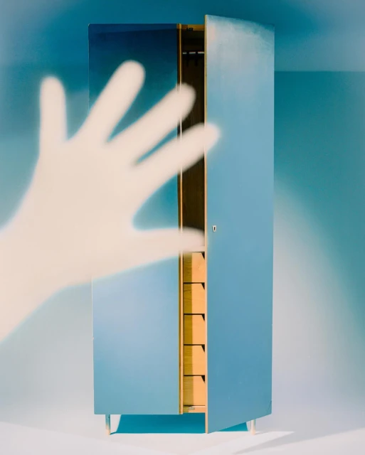 Minimalist photograph of an open, light blue armoire with natural wood interior, next to a ghostly hand shadow on a blue wall.