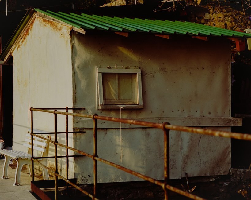 A quaint, slightly weathered shed with a green roof stands near a rusty metal railing and a white bench, with natural light highlighting its textures.