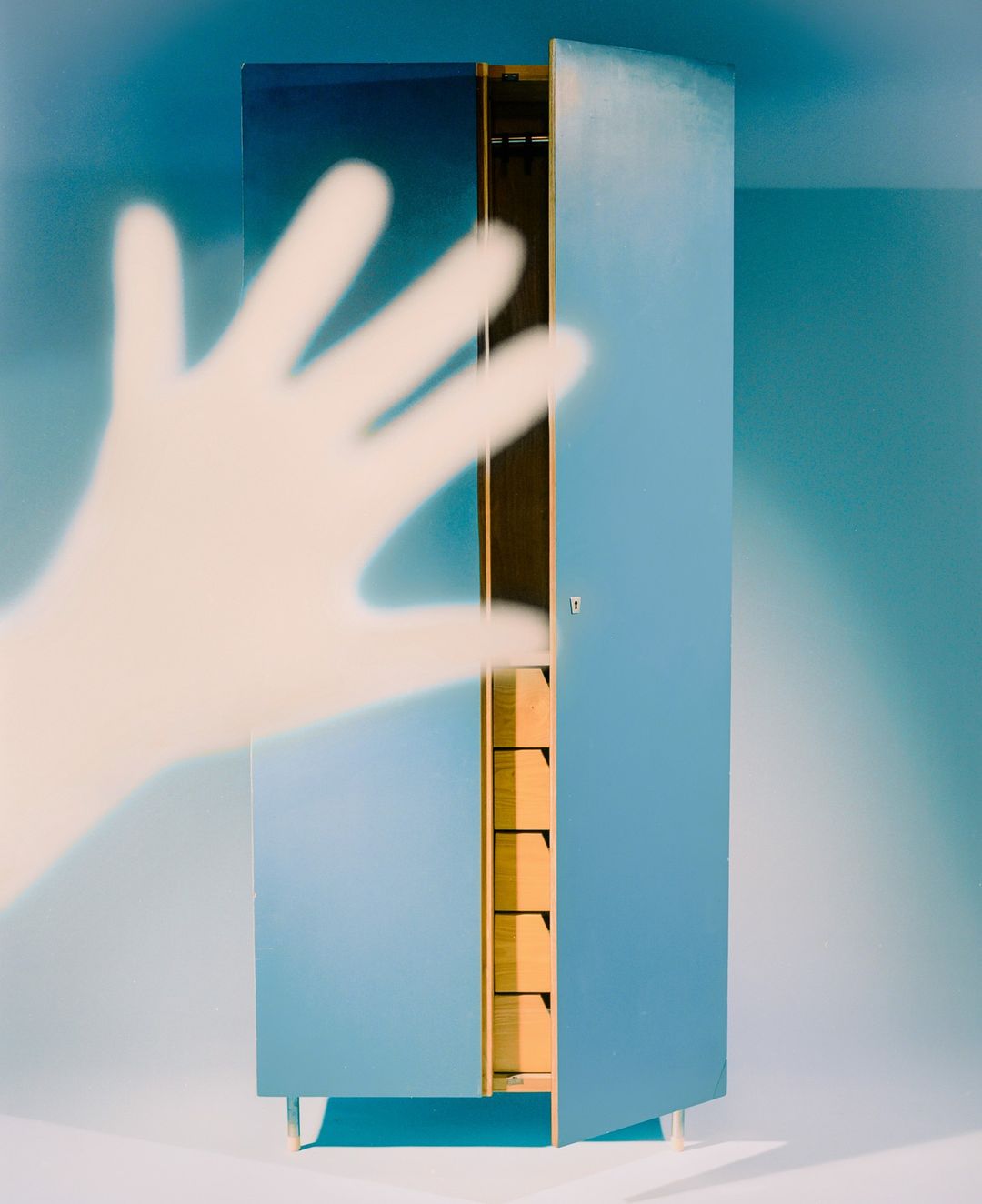 Minimalist image of a blue wardrobe with open door, partially visible hand, against a pale blue backdrop in a studio setting.