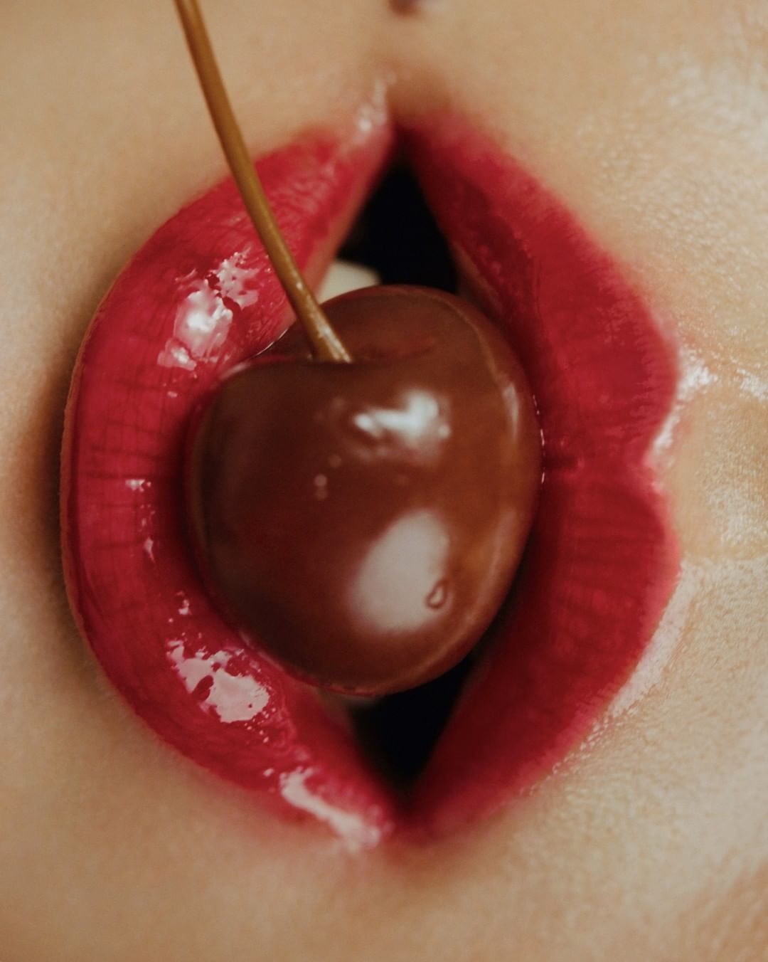 Close-up of a woman's mouth with red glossy lips and a cherry held in her mouth, creating a sensual and artistic beauty image.