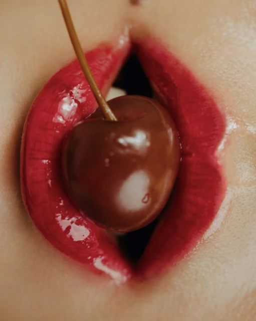 Close-up of a woman's mouth with red glossy lips and a cherry held in her mouth, creating a sensual and artistic beauty image.