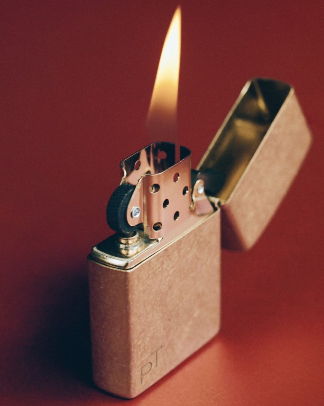 Brass Zippo lighter with flame on red background. The lighter is partially open, showing the mechanics. Warm light reflects on the surface.