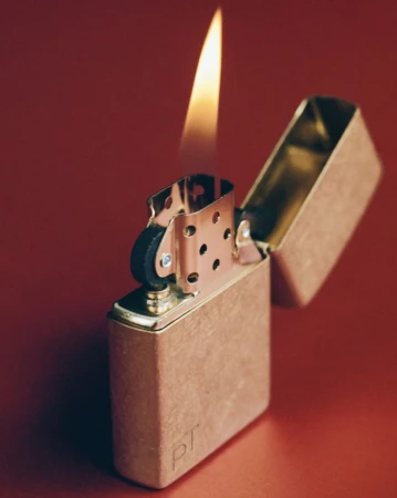 Brass Zippo lighter with flame on red background. The lighter is partially open, showing the mechanics. Warm light reflects on the surface.