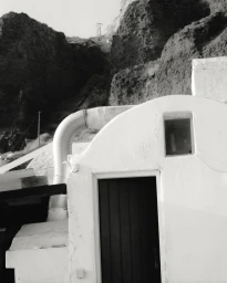 Monochrome architectural photograph of a white building with a dark door set against a rugged rocky cliffside.