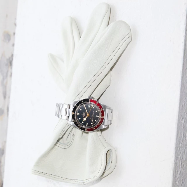 Wristwatch with a stainless steel bracelet displayed on a folded white leather glove against a textured white surface. Timepiece details.