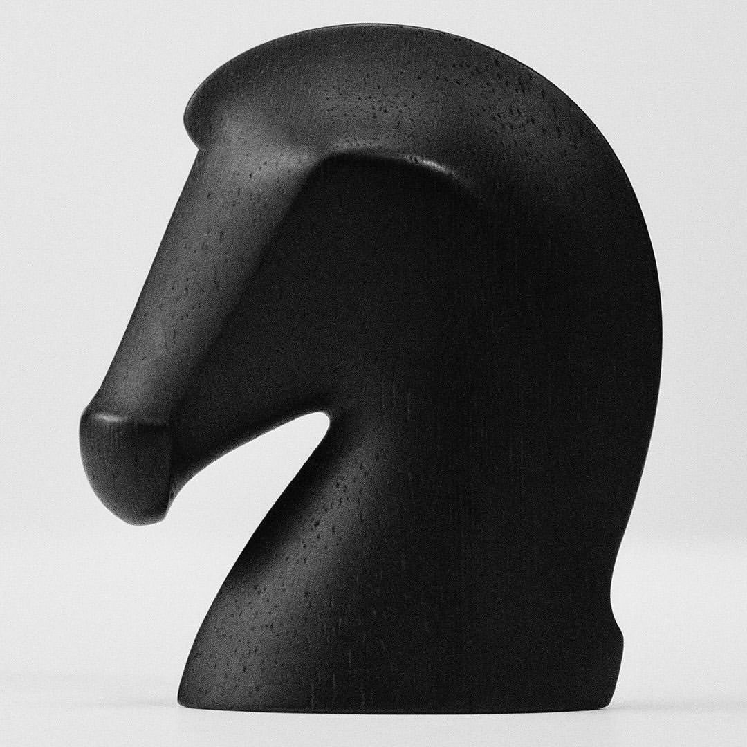 Close-up of a black wooden chess piece in the form of a horse head, set against a soft gray background. Smooth texture is visible.