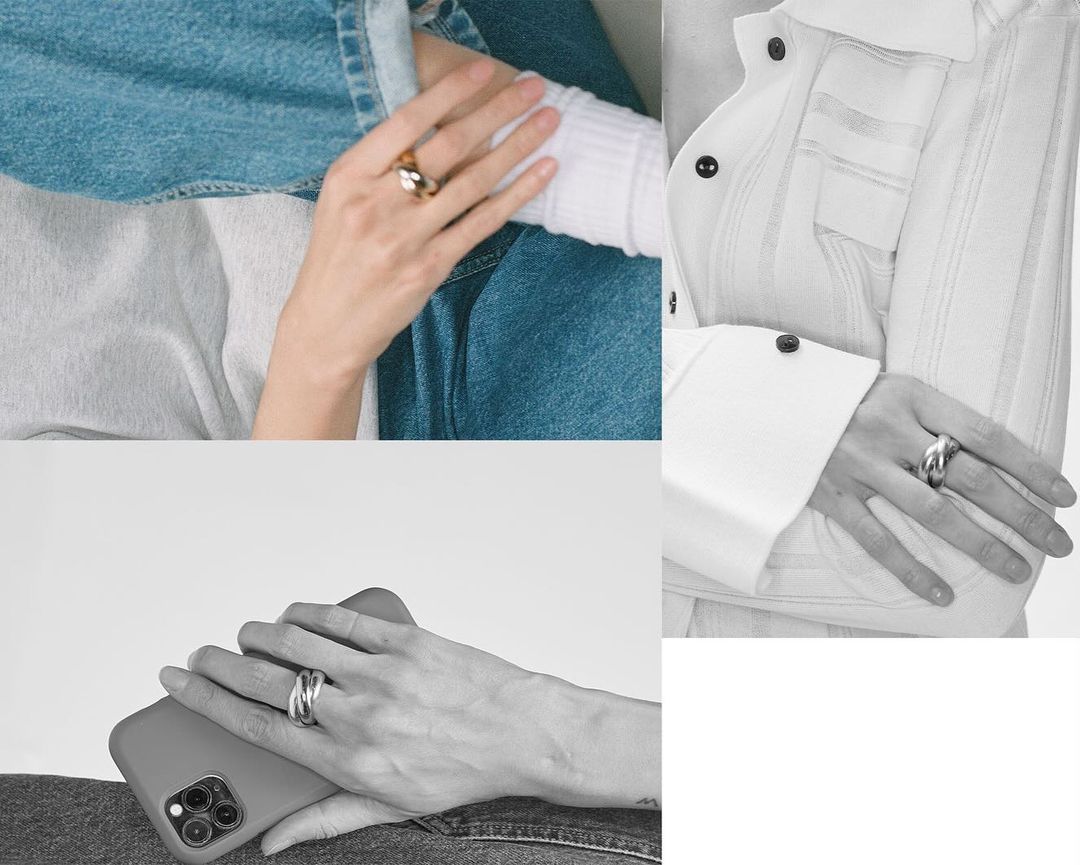 Close-up of hands wearing gold and silver rings, featuring denim and white striped shirt sleeves in a minimalist, indoor fashion setting.