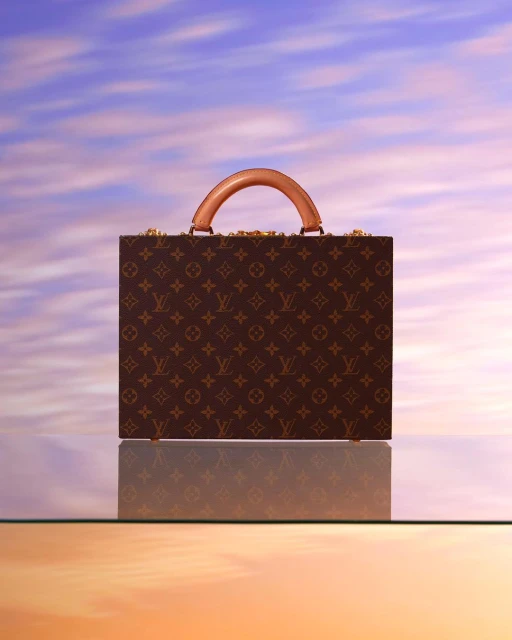 Luxury brown Louis Vuitton trunk on a glass surface reflecting a sky background with wispy clouds and pink and orange hues.