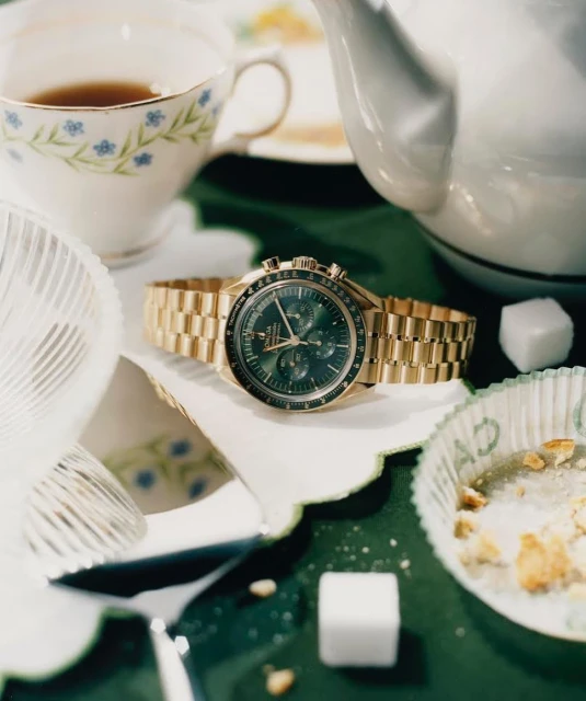 Gold Omega Speedmaster watch on a table with a tea cup, tea pot, sugar cubes and a cupcake wrapper on a green background.