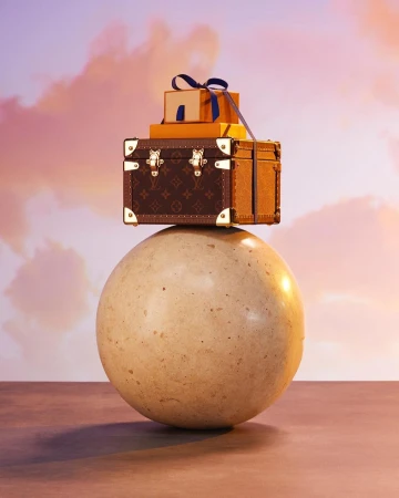 Louis Vuitton trunk and gift boxes stacked on a tan sphere against a pink and lavender cloudy sky background in studio setting.