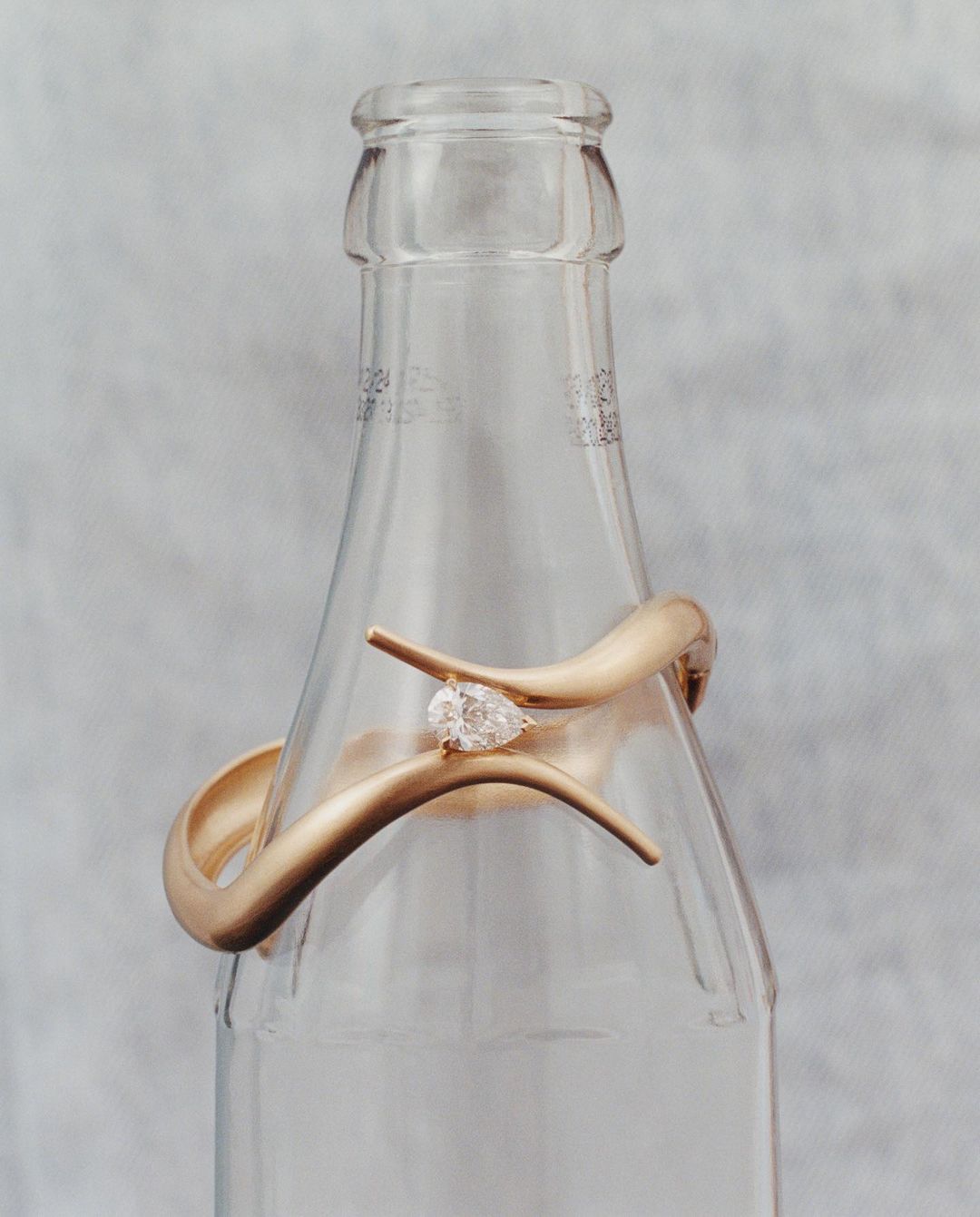 A gold bracelet with a pear-shaped diamond rests on a clear glass bottle against a neutral background, showcasing a modern jewelry design.