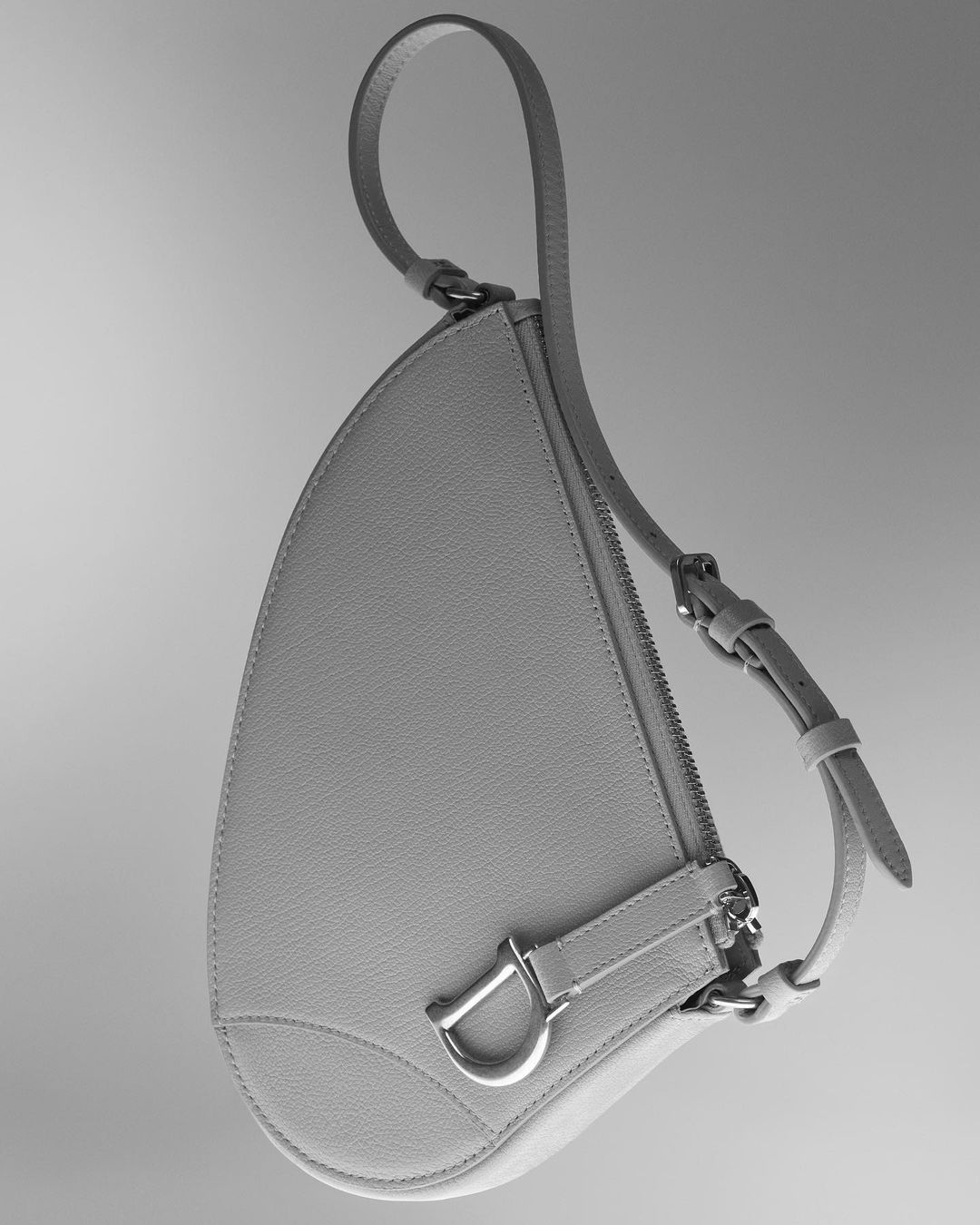 Studio shot of a gray Dior Saddle bag with silver hardware and a leather strap against a neutral background, showcasing its unique shape.