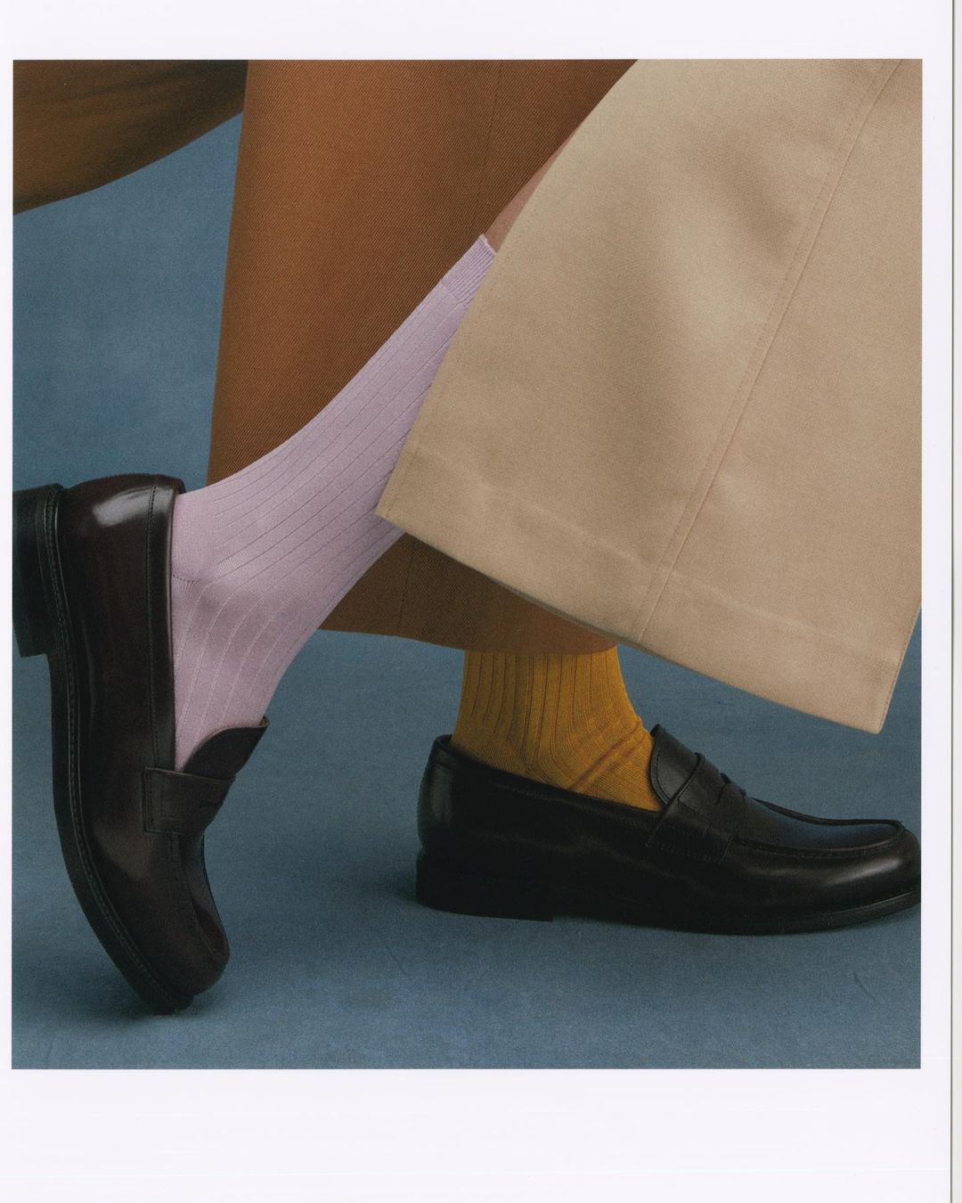 Close-up shot of two legs wearing socks and shiny black loafers on a blue surface, showcasing a fashion footwear concept.