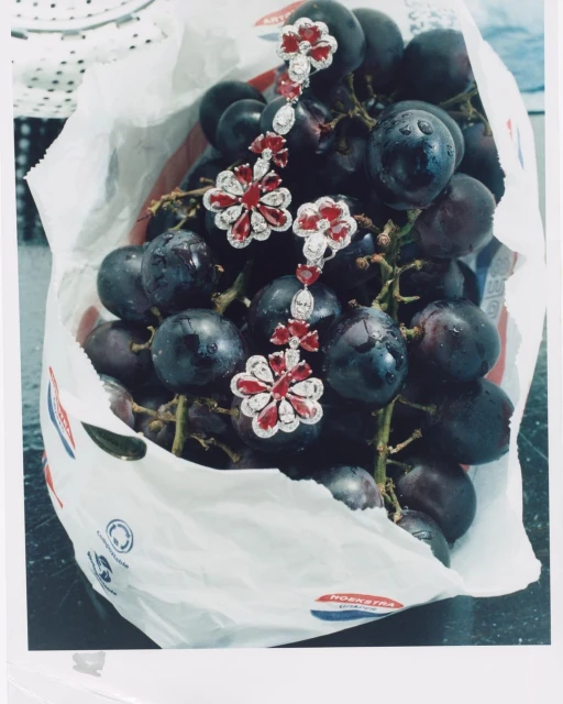 Elegant flower-shaped earrings with rubies displayed atop a pile of fresh, dark purple grapes in a white paper bag.