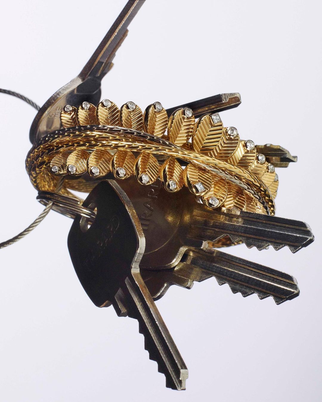 Close-up photo of a cluster of silver keys connected to a gold-colored ornamental leaf and metal band on a white background.