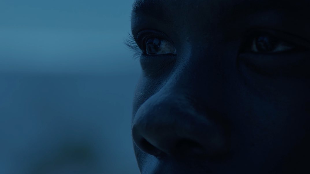 Close-up of a young Black adult's face, with focus on their eyes, eyelashes, and skin texture under dim blue lighting.