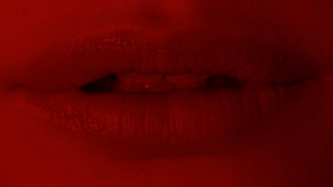 Macro view of open human mouth with red lipstick and red lighting creating an intense, sensual close-up in a vibrant, monochromatic color scheme.