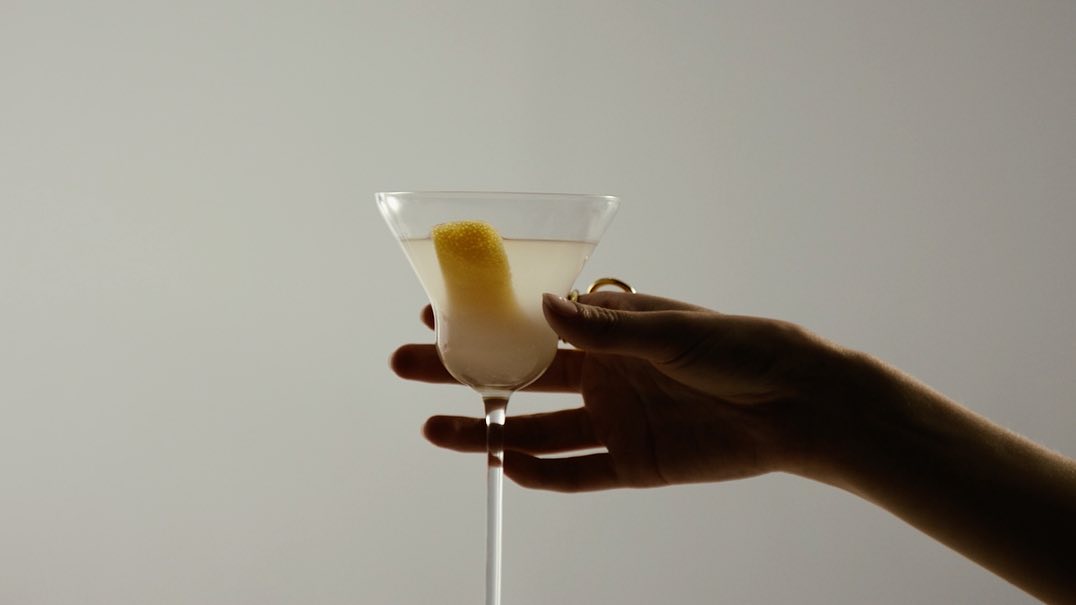 Close-up of a martini glass with a lemon twist held by a hand against a plain background. The cocktail is pale in color.