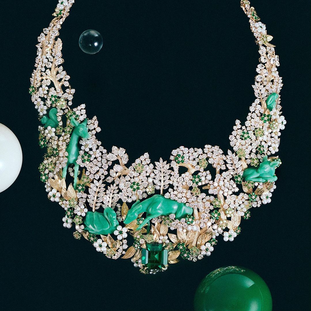 Elaborate necklace featuring diamonds, green gems, gold branches, and carved jade animal figures set against a dark background.