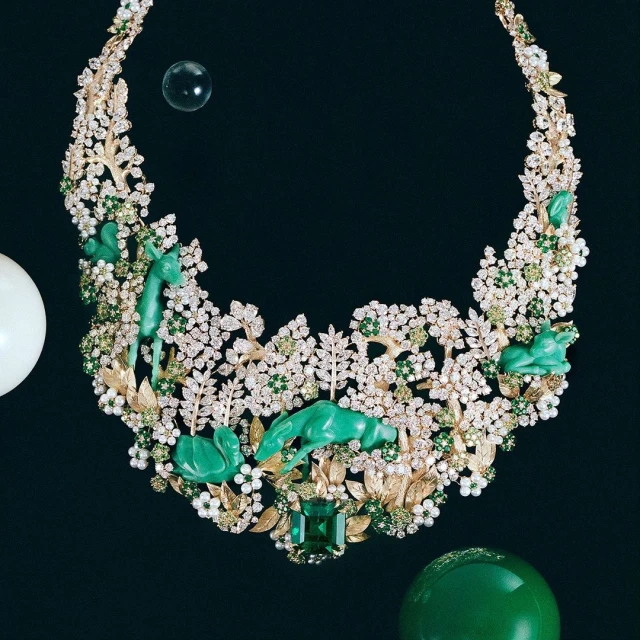 Elaborate necklace featuring diamonds, green gems, gold branches, and carved jade animal figures set against a dark background.