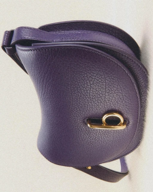 Close-up of a stylish saddle bag in vibrant purple leather with gold hardware accents, set against a plain, light-colored background.