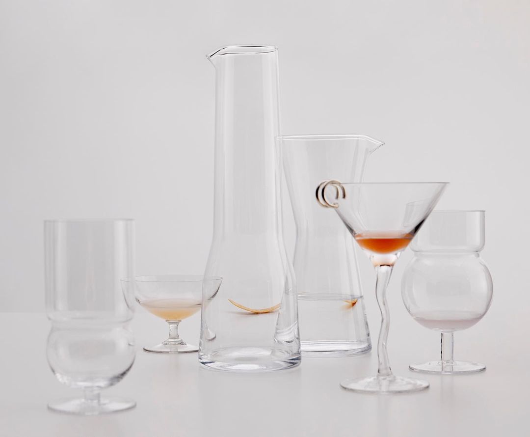 Minimalist still life features an arrangement of five clear glass vessels in various shapes and sizes against a white background.
